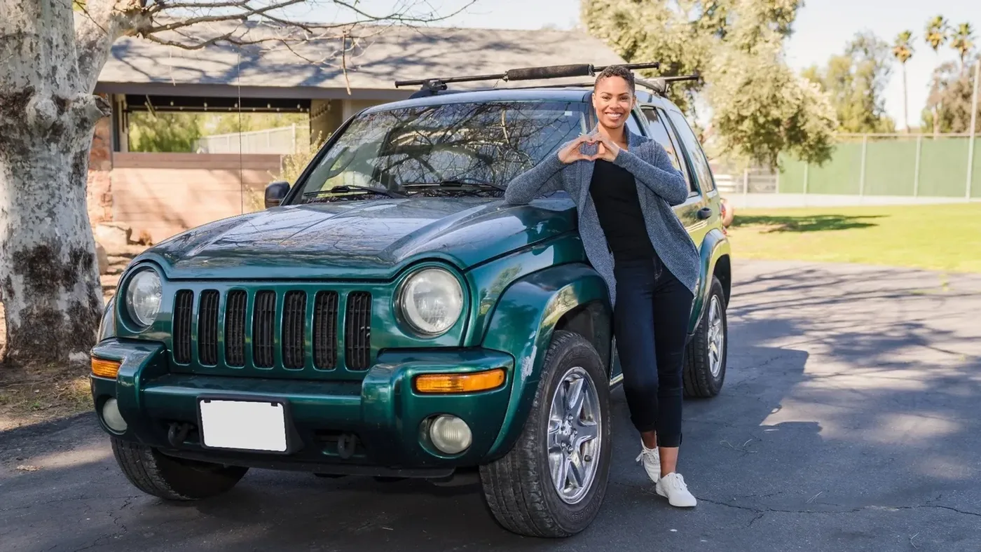 A smiling woman leaning on a green SUV, making a heart shape with her hands.
