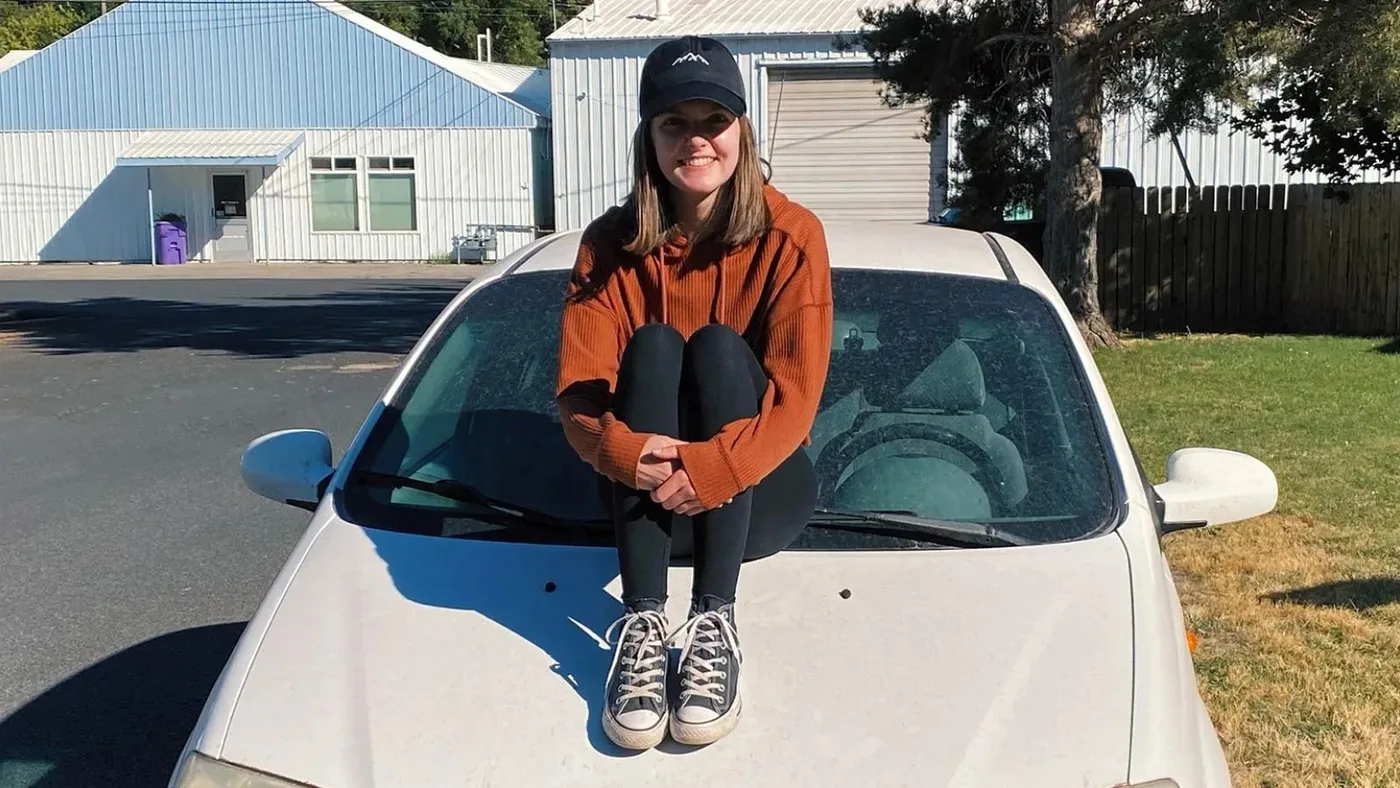 A smiling young woman in a cap and rust sweater sitting on the hood of a white car.