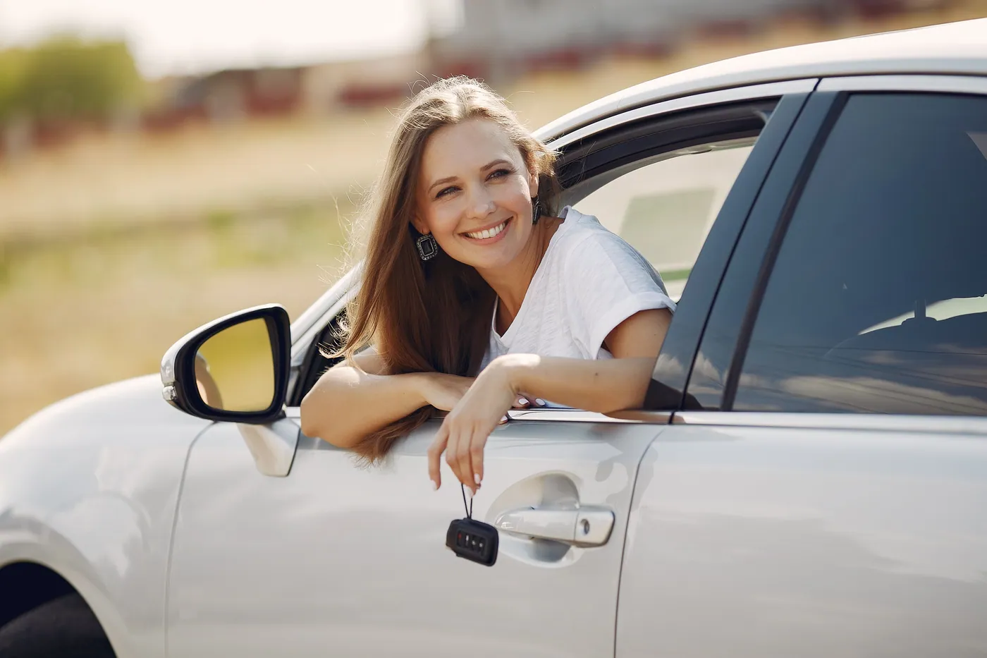 A smiling woman leaning out of the window of her white car, holding the key.